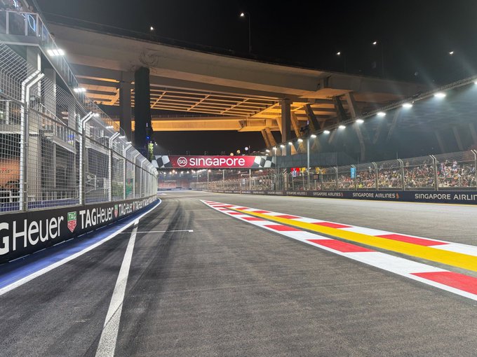 A nighttime view of the Singapore street circuit track with visible 'Singapore', 'TAG Heuer', and 'Singapore Airlines' signage around the race barriers.