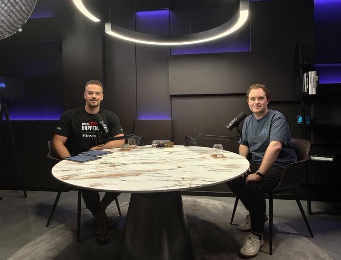 Two men sit at a round marble table in a modern studio with dramatic overhead lighting and purple accents, recording what appears to be a podcast, with one wearing a shirt reading "WE ARE ALL HAPPEN." The setting matches the accompanying post about a long-form conversation with Lucas, Founder and CEO of Variational, who co-founded a hedge fund acquired by Digital Currency Group before building Variational.