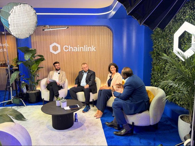 Four people in business attire sit in white armchairs in a studio with blue walls and green plants, having a panel discussion at a 'Chainlink' branded event.
