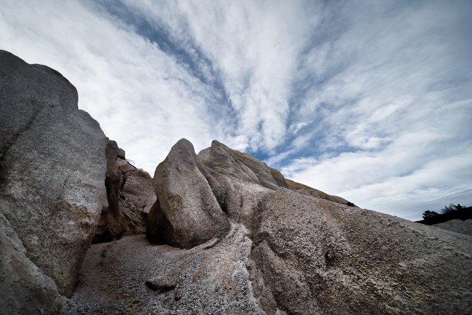 A dramatic low-angle photograph of a large, rugged rock formation set against a bright blue sky filled with scattered white clouds.