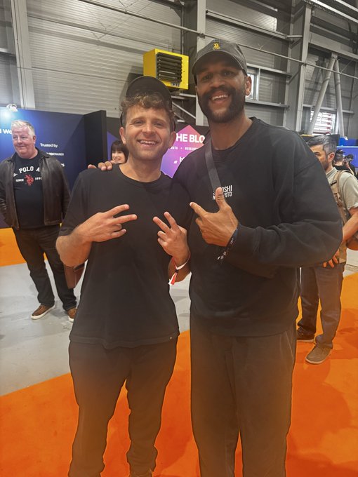 The image shows two men standing next to each other, both smiling at the camera. One of them has his hand raised in a peace sign gesture. The background reveals that they are inside an exhibition hall or a convention center, with several people present around them. There is also a yellow object visible in the scene.
