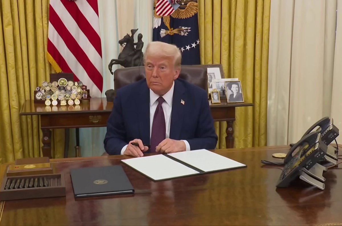 The image shows a man sitting at a desk, wearing a suit and tie, with his hands on a piece of paper. The setting appears to be an office or a room within an office building, as there are multiple chairs present around the scene. A TV is also visible in the background. The post includes text that reads "BREAKING BOOOM NEWS WHO'S F*KING READY FOR TRUMP WILL SIGN AN URGENT EXECUTIVE ORDER TODAY AT 3:30 PM ET, RIGHT BEFORE MARKETS CLOSE! HE NEVER SIGNS BEFORE MARKETS CLOSE UNLESS SOMETHING BULLISH IS HAPPENING". The image seems to be related to a political event or situation that is about to take place.