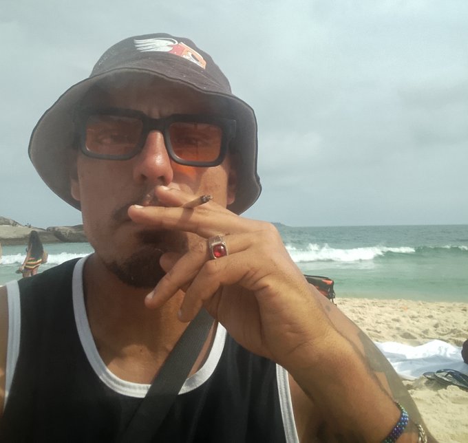 A man with a goatee, wearing a bucket hat with a flag logo and orange-tinted sunglasses, smokes a cigarette on a beach while wearing a red gemstone ring.