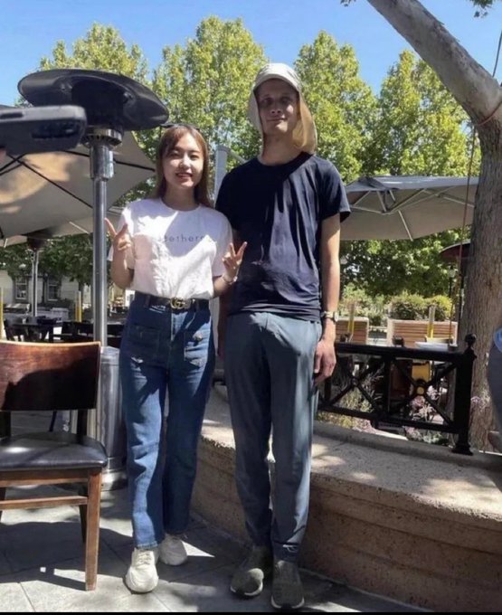 A young woman in a white t-shirt printed with "Ethereum" poses next to a tall man in a black shirt, both flashing peace signs at an outdoor café. The image illustrates the post's tongue-in-cheek claim about uncovering an "alt" identity through social media analysis, with the woman's Ethereum shirt serving as the visual punchline connecting the two.