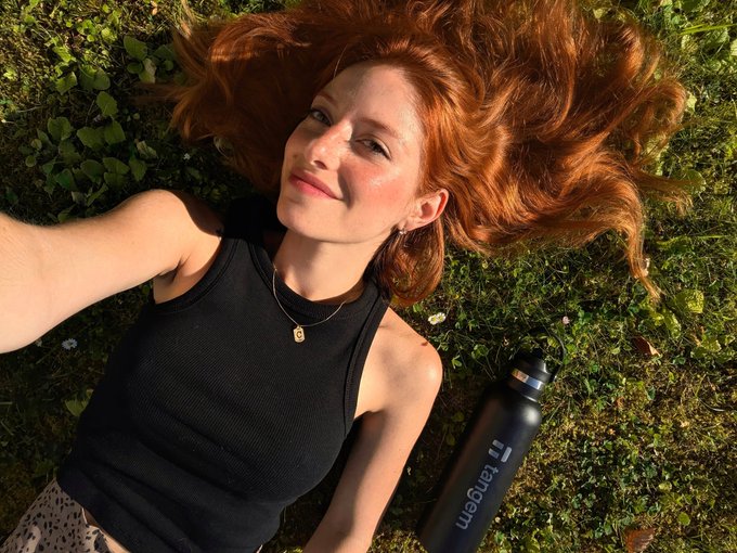 A woman with red hair lying on green grass in a black tank top, posing for a selfie next to a black 'Usain' branded water bottle.