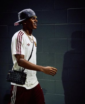 A young man in a white Adidas shirt and maroon pants with a gray cap and crossbody bag walks along a wall in dim lighting.