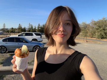 A woman with short brown hair holds up a small taco wrapped in paper while taking a selfie in an outdoor parking lot, playfully referencing the post's question about choosing between a BDR at Corgi or being a goat in Sonoma by posing as if enjoying a goat-related snack.