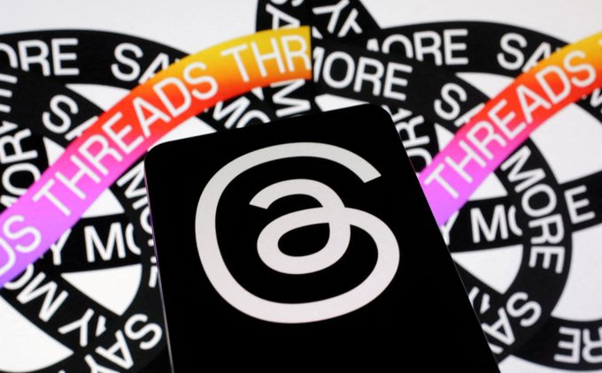 The image features a black cell phone with a white "a" logo, placed against a background of colorful text and shapes. The text includes phrases such as "NEW: $META Threads said it has now reached 500M monthly users." This suggests that the cell phone is associated with a popular social media platform or brand.
