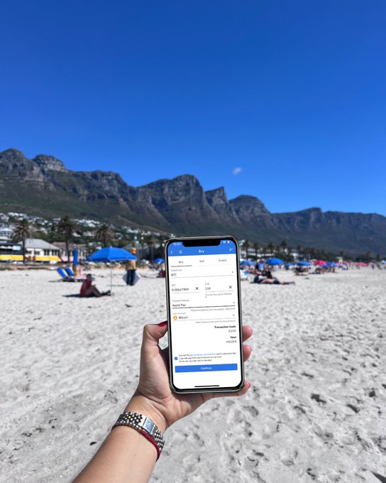 A hand holds a smartphone displaying a currency conversion app at a beach with mountains in the background, showing exchange rates between USD and ZAR.
