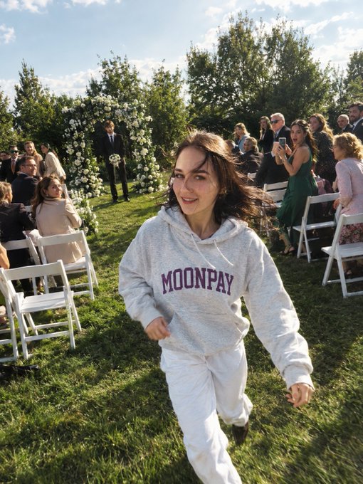 A young woman in a gray 'MOONPAY' hoodie running playfully across a lawn at an outdoor wedding ceremony.