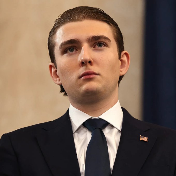 A young man in a dark suit, white shirt, and navy tie with an American flag lapel pin looks upward with a serious expression.