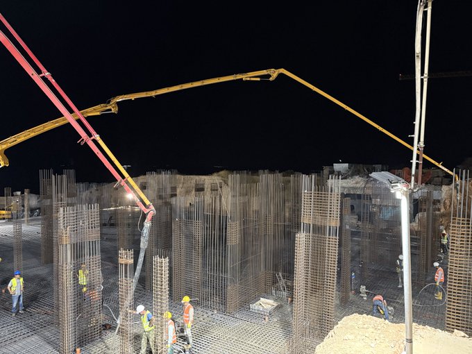 Construction workers in hard hats and high-visibility vests work at night pouring concrete at a building site with rebar columns and concrete pump booms visible.