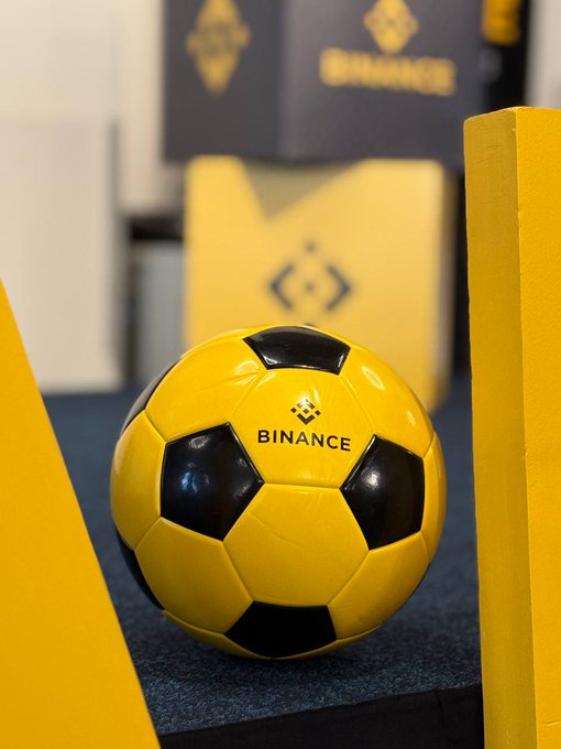 The image features a yellow soccer ball with black spots, which appears to be a Binance wallet. The soccer ball is placed near some boxes or crates, possibly representing an office setting. The background of the image includes a white wall and a gray box, creating a contrast between the vibrant colors of the soccer ball and the more neutral tones of the surroundings.