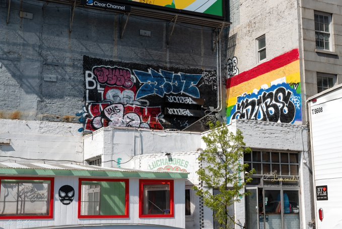 An urban street scene featuring a building wall covered in colorful graffiti and a rainbow mural, with a Clear Channel billboard above, red-trimmed storefronts, and a tree in the foreground.