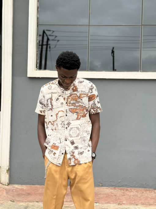 A young man looks down while wearing a white shirt printed with vintage nautical map designs paired with mustard yellow pants, standing outside a gray building with large windows. The image appears unrelated to the post, as it shows a casual streetwear fashion shot rather than anything visually connected to the AI Assistant feature being promoted on @Scallop_io.