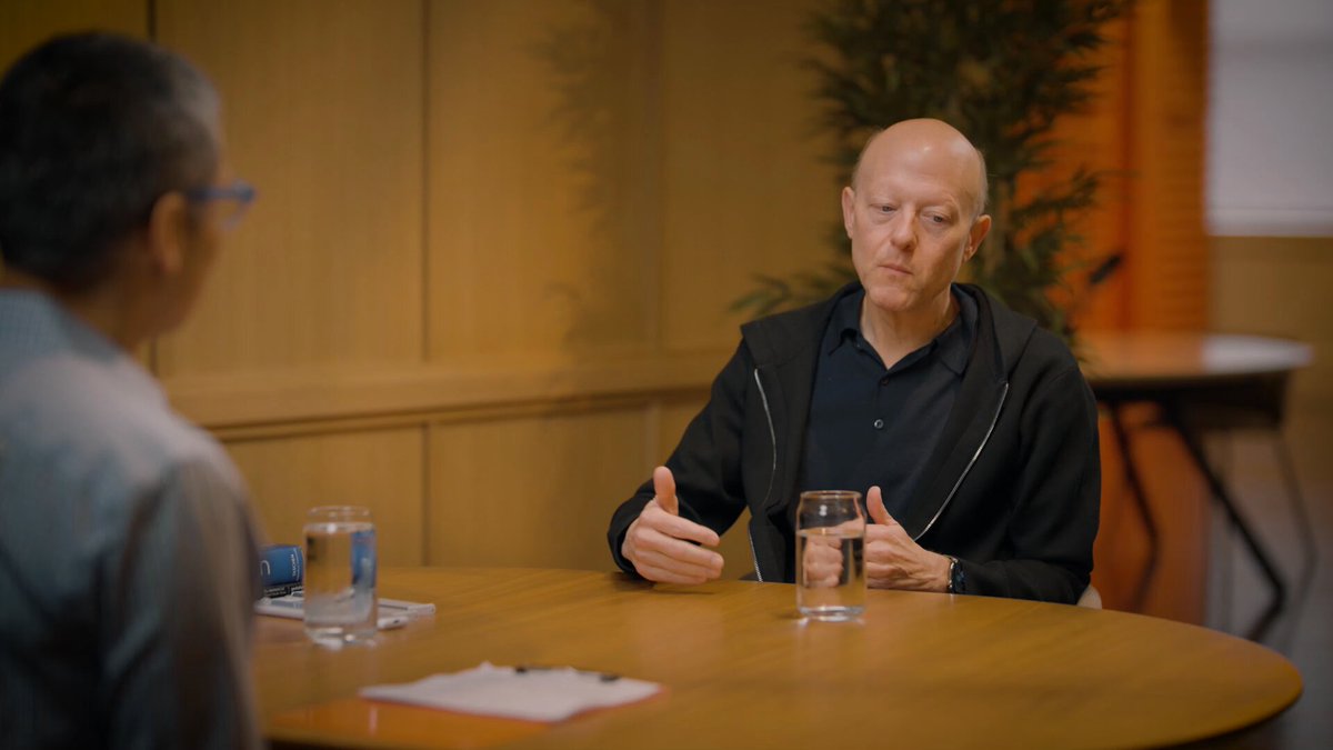 The image shows a man sitting at a table, giving a thumbs up gesture while holding a glass of water. The scene takes place in an office setting with wooden walls. There are two other people present in the room, one standing behind the seated man and another person partially visible on the right side of the frame. The overall atmosphere suggests that they might be discussing or reviewing something related to financial infrastructure, as indicated by a post on @ycombinator about regulatory clarity and its impact on various use cases across different asset classes.