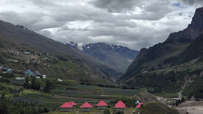 A mountain valley landscape with green terraced fields, small houses with red roofs in the foreground, and snow-capped peaks under a cloudy sky.