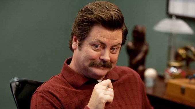 A man with a mustache in a red checkered shirt making a smug expression while sitting at a desk.