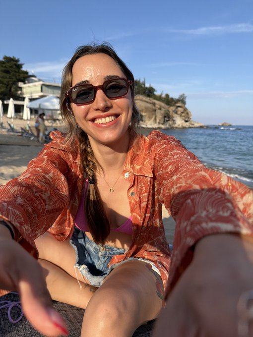 A smiling young woman with sunglasses and a braided hairstyle taking a selfie at the beach, wearing an orange patterned shirt over a bikini top with denim shorts.