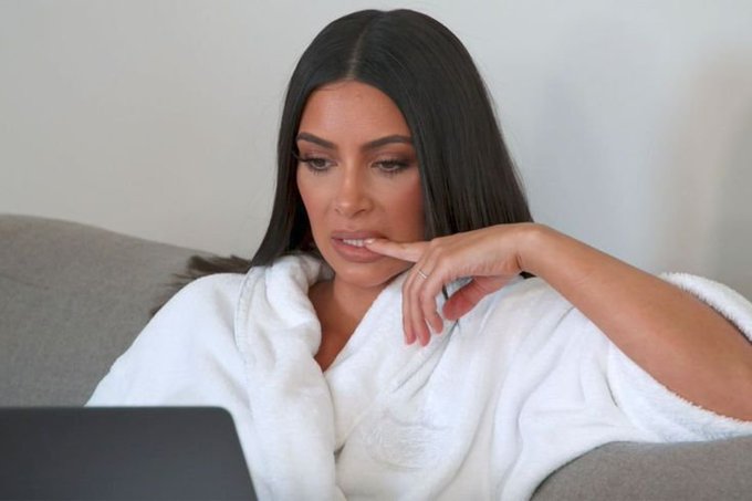 A woman with long dark hair, wearing a white robe, sits on a couch while looking thoughtfully at a laptop screen.