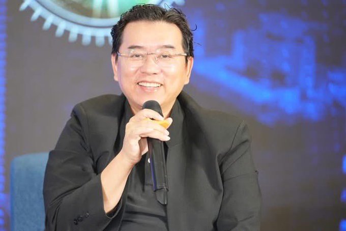 An older Asian man wearing glasses and a black blazer smiling while holding a microphone, appearing to be speaking at a conference or event with a blue technical background.