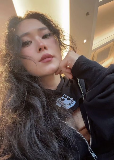 A close-up selfie of a young woman with long dark hair wearing a black hoodie with a small smiley face patch, posing against a softly lit background.