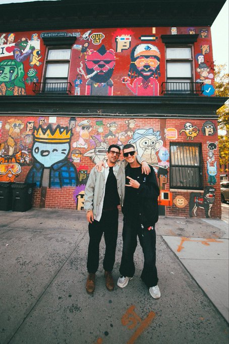 Two people pose together in front of a brick building covered in colorful street art and cartoon-style murals.