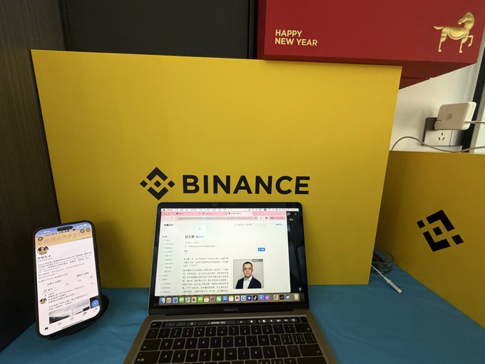 A desk setup featuring a laptop and smartphone in front of a yellow 'BINANCE' sign, with a red 'HAPPY NEW YEAR' banner displaying a gold horse above it.