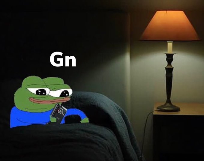 The image features a cartoon frog sitting on a bed, holding a cell phone with its left hand while looking at something else with its right hand. The background of the image shows a lamp that appears to be turned off or dimmed, creating a cozy atmosphere in the room. The text "GN" is written above and below the frog's head, which might be related to the post about the Good Night network.