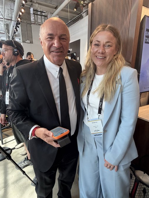 A man in a black suit and tie holding a smartphone stands next to a smiling blonde woman wearing a light blue blazer and a 'SPEAKER' lanyard badge at what appears to be a conference or event.