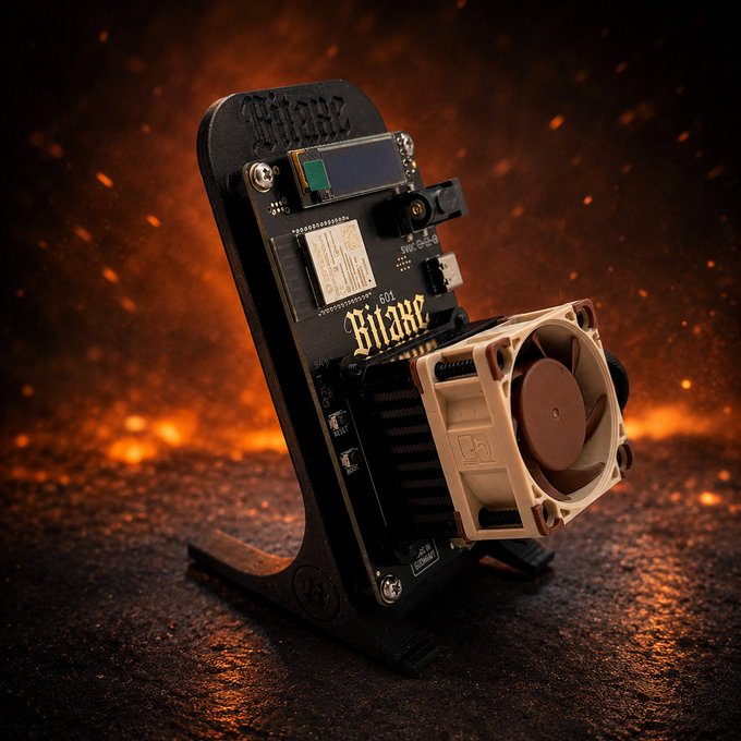A DIY electronics device named 'Bitaxe' stands upright, featuring a circuit board, small screen, camera module, and a cooling fan with a Noctua-style logo, displayed against a fiery, ember-filled background.