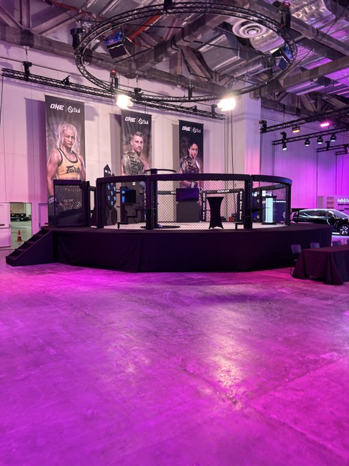 An empty MMA octagon set up in a large venue, illuminated by purple lighting. Banners for 'ONE' featuring fighters stand behind the cage, and a black car is visible in the background.