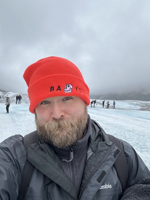 A selfie of a bearded man wearing a red beanie with "BABY" and a cartoon face logo, and a gray Columbia jacket, taken on a glacier with other tourists in the background.