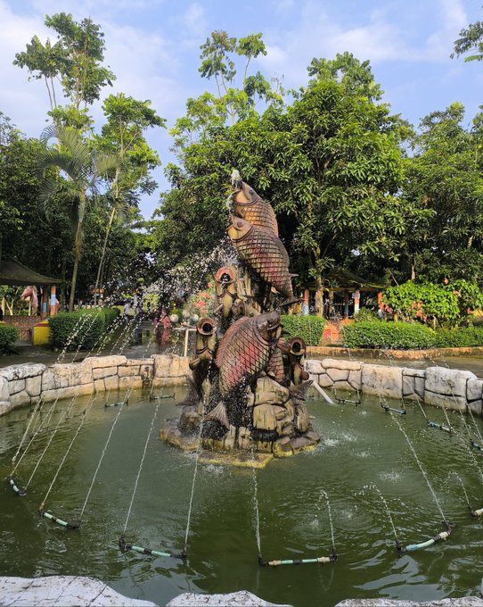 The image features a large fountain surrounded by trees, creating an outdoor scene that appears to be located within a park or garden setting. The water from the fountain is spraying up into the air, adding movement and life to the scene. In the background, there are several people enjoying their time in the area, possibly admiring the beautiful surroundings or engaging in other activities. The image also includes text elements such as "Good Morning All My X Family GM Happy Wednesday with @NomismaNetwork & @useTria 3look".