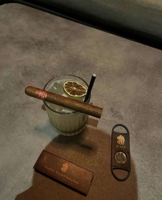 A drink with a lemon slice and a cigar resting on its rim sits next to an 'EMS' branded cigar cutter and an 'English Market Selection' cigar box on a concrete surface.