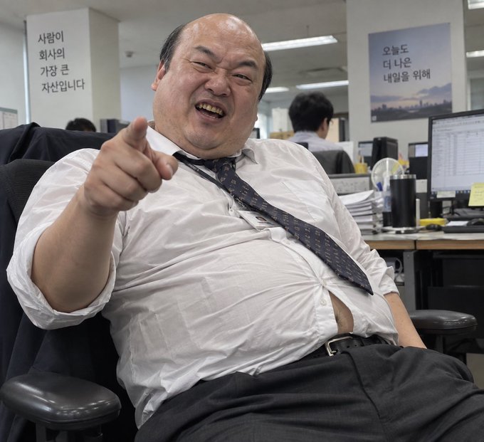 A photo of a large man in a white shirt and tie, laughing and pointing at the camera in an office setting with Korean text on signs in the background.