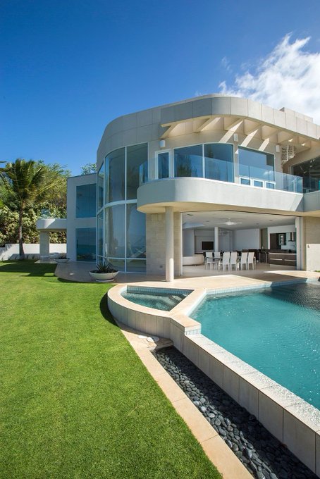 A modern, luxurious two-story home features curved architecture, large glass windows, a balcony, and an infinity pool overlooking a well-manicured lawn.