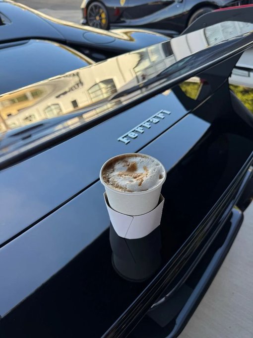 A small paper cup of coffee placed on the rear decklid of a black Ferrari, with another sports car visible in the background.