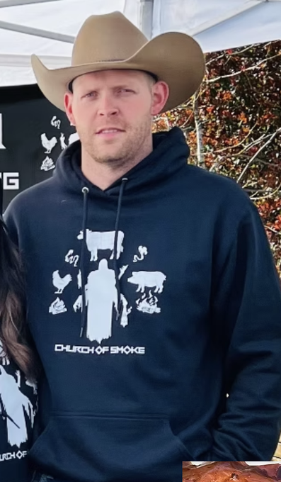 A man wearing a tan cowboy hat and a dark hoodie with 'CHURCH OF SMOKE' printed on it, standing at an outdoor event.