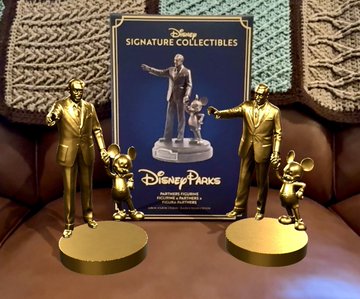 Two gold Disney Parks statues of Walt Disney with Mickey Mouse flank a 'Signature Collectibles' box on a brown leather chair.