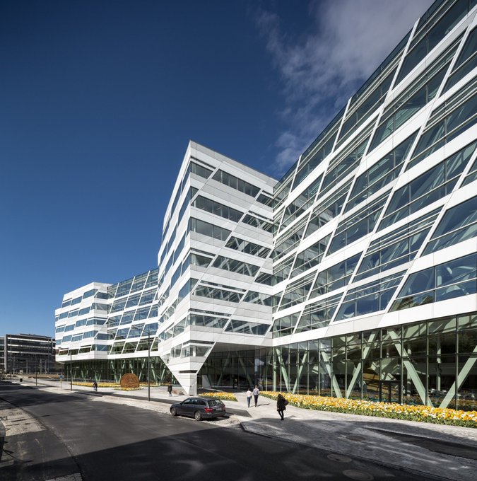 A modern, multi-story glass office building with a distinctive angular facade stands under a bright blue sky, with a few pedestrians walking near the entrance. The image likely represents the corporate headquarters or a regional office building related to Swedbank AB's recent investment activity in Strategy.