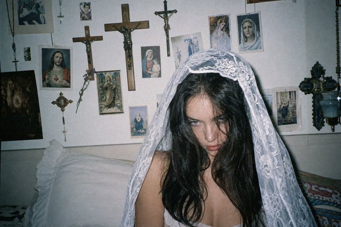 A moody portrait features a woman wearing a white lace veil, with crucifixes and religious images adorning the wall behind her.
