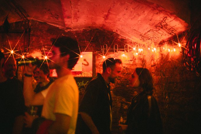 The image shows a group of people standing around each other, with one person holding a drink. They are gathered inside an underground room that has neon lights hanging from the ceiling, creating a vibrant atmosphere. The photo appears to have been taken at night or during low light conditions, as it is described as being "very dark" and "very dim". The image also contains text that reads: "A moment in the techno dungeon. The @Zcash Blockspäti opening went off last night." This suggests that there might be a special event happening inside the room at this time or that the neon lights are part of an ongoing celebration.