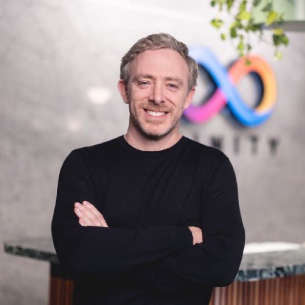 Photo of a smiling man with short blonde hair and beard, wearing a black long-sleeve shirt with arms crossed, standing in front of a colorful infinity logo.