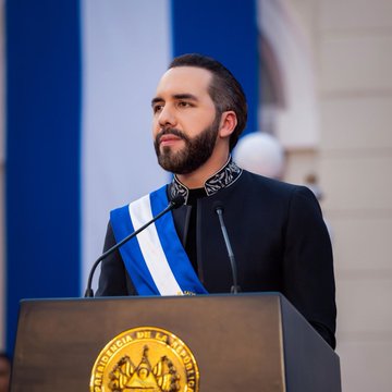 A man with a beard wearing a sash with El Salvador's colors speaks at a podium bearing the seal "Casa Presidencial de El Salvador," appearing to be President Nayib Bukele. The image accompanies news of El Salvador expanding its Strategic Bitcoin Reserve to nearly 7,800 BTC, reinforcing the country's continued commitment to its daily Bitcoin purchasing strategy.