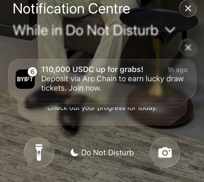 The screenshot shows an Apple Vision Pro notification center in Do Not Disturb mode displaying a promotional Bybit notification reading "110,000 USDC up for grabs! Deposit via Arc Chain to earn lucky draw tickets. Join now." This relates to the post by highlighting Bybit's aggressive push notifications soliciting cryptocurrency deposits with promotional offers.