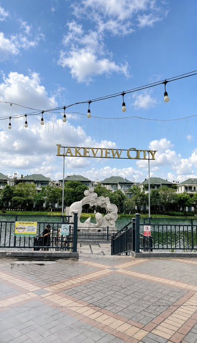 An outdoor daytime photo of the entrance to Lakeview City, featuring a decorative gate with a white sculpture, a sign, string lights overhead, and a paved walkway.