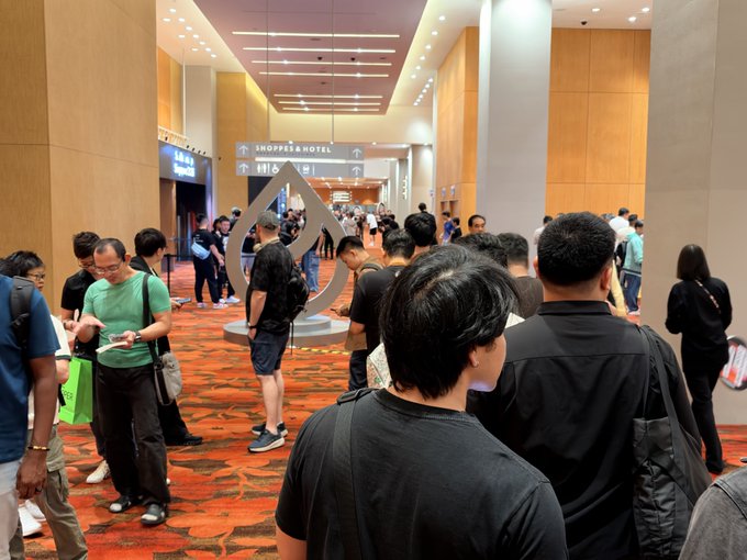 A crowded hallway inside a shopping mall or hotel convention area with a patterned carpet, where attendees are gathered near signs for 'Shops & Hotel' and a large metal sculpture.