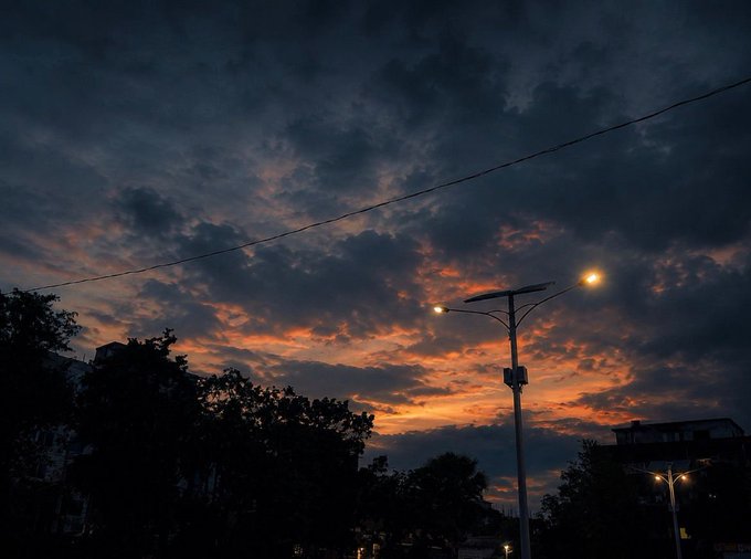 A dramatic dusk photograph of a cloudy sky tinged with orange sunset light, featuring a silhouetted street lamp and tree line in the foreground.