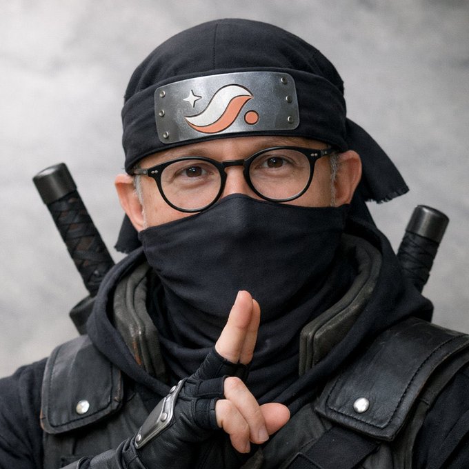 Person dressed as a ninja in a black outfit with headband, mask, and dual swords on their back, holding up two fingers in a peace sign.